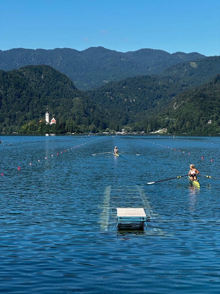 Ruderer ziehen bei einer Regatta ihre Bahnen auf dem spiegelglatten Bleder See, dahinter die Insel mit der Kirche.