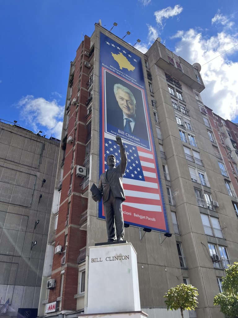 Bill-Clinton-Statue vor einem Hochhaus mit Porträt-Banner und US-Flaggen in Priština
