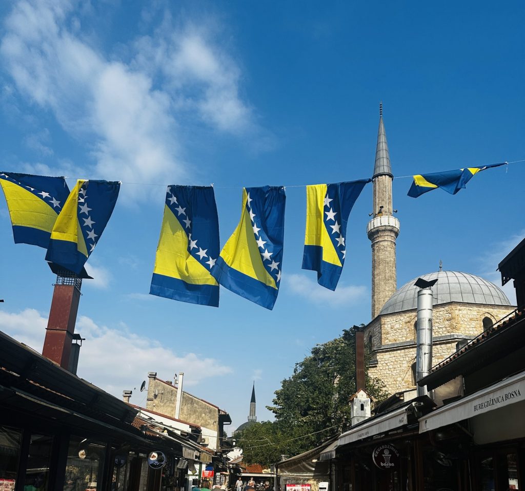 Bosnische Fahnen spannen sich über eine Gasse der Baščaršija, dahinter Minarett und Moscheekuppel.
