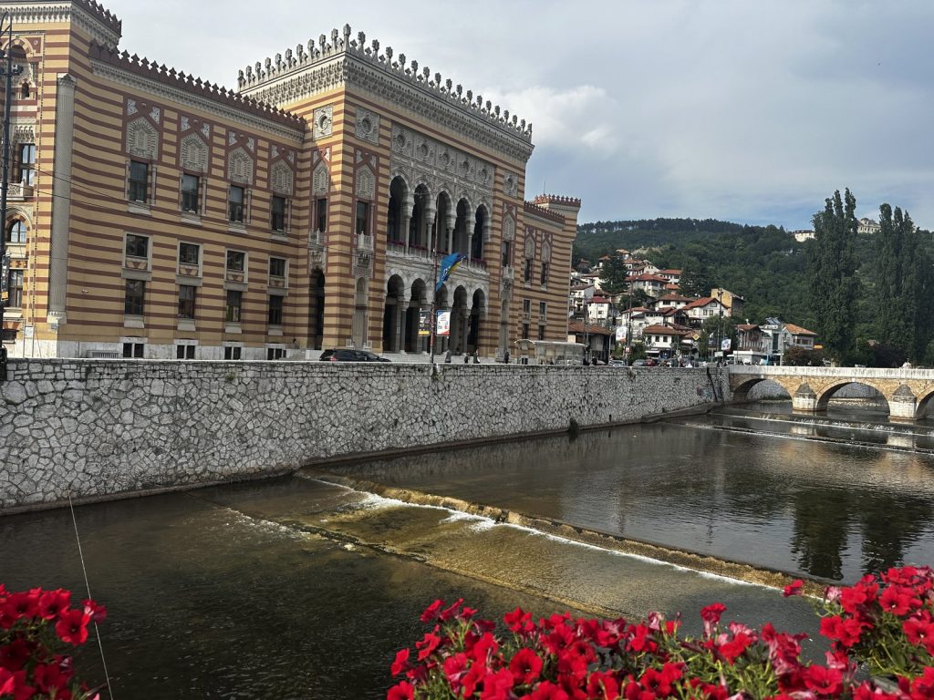Das gelb-braun gestreifte Rathaus Vijećnica am Fluss Miljacka, davor eine Reihe roter Blumen.