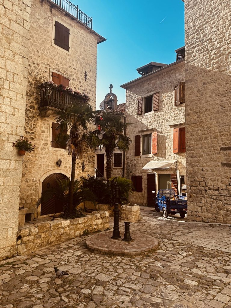 Ein stiller, sonniger kleiner Platz in der Altstadt von Kotor mit Kopfsteinpflaster, Palme, altem Brunnen und honigfarbenen Steinhäusern.