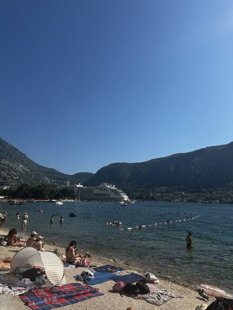 Schmaler Strand an der Bucht von Kotor mit einem Kreuzfahrtschiff im Hintergrund.