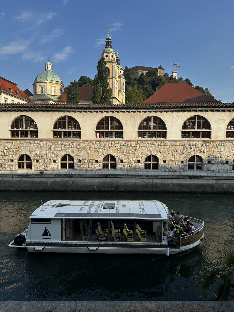 Die Markthalle am Fluss Ljubljanica mit Ausflugsboot und der Domkuppel dahinter.