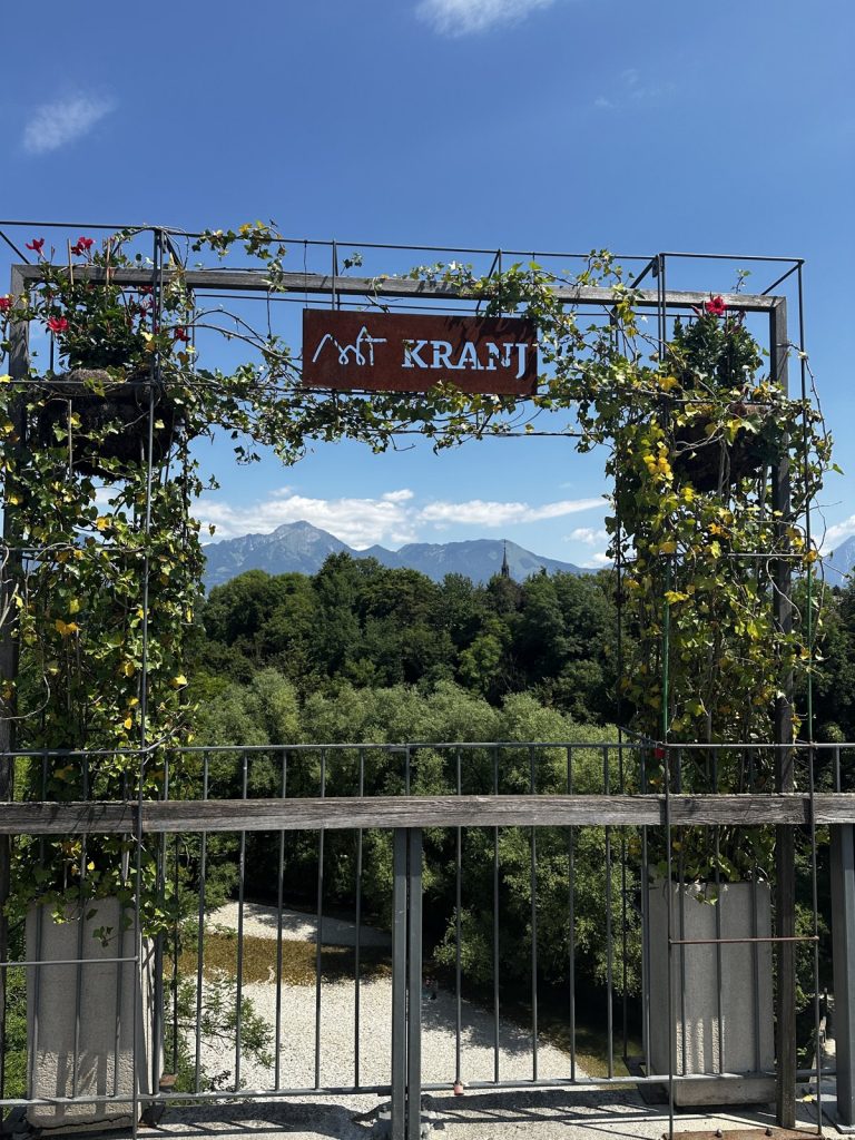 Das „KRANJ"-Torschild mit Weinranken und den Bergen im Hintergrund.